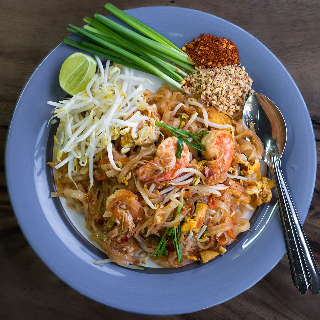 Prawn pad thai with flat rice noodles and egg, beside bean sprouts, chives, a lime wedge, chili flakes and crushed peanuts