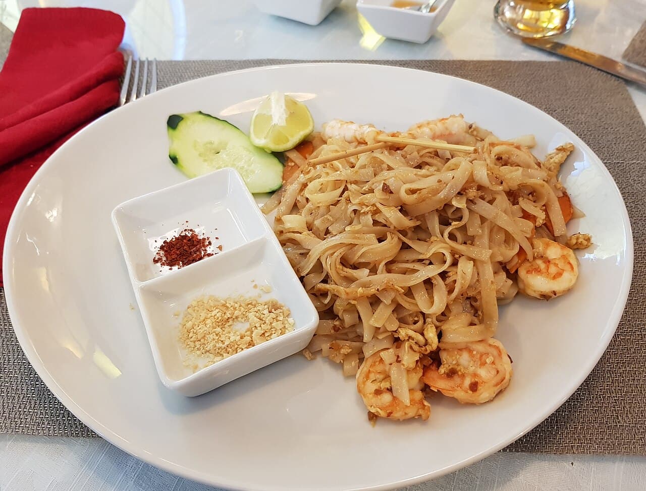 A plate of pad thai with shrimp, a lime wedge and cucumber, beside small dishes of chili flakes and crushed peanuts