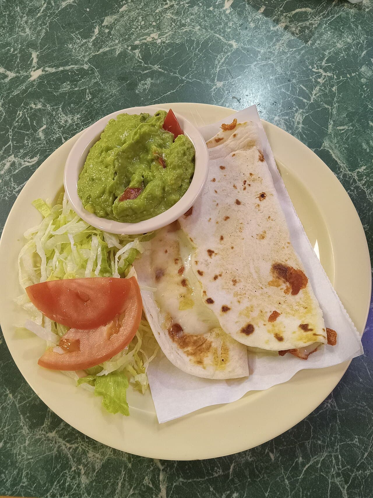 A plate with a bowl of guacamole, two quesadillas, shredded lettuce and tomato slices
